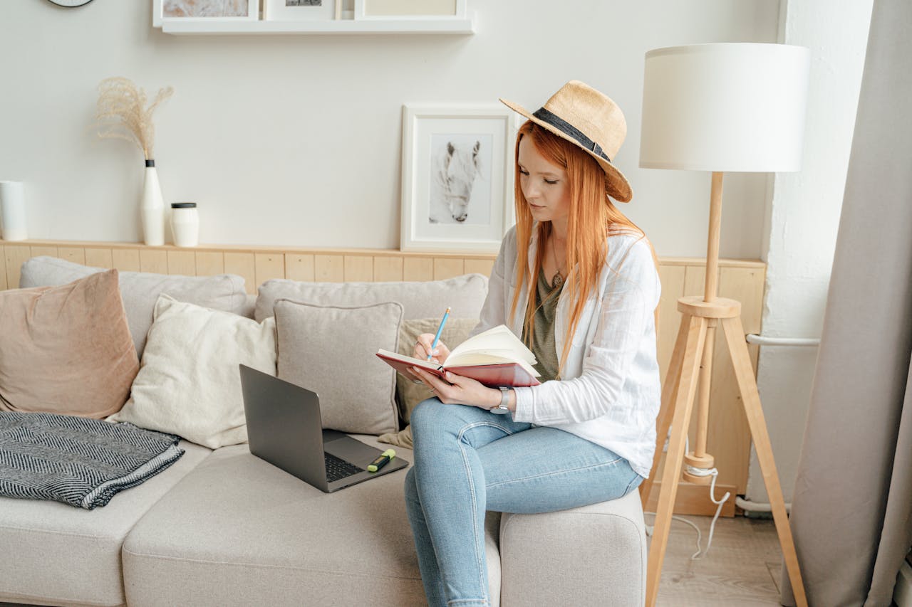 Woman sitting on sofa, writing in notebook with a laptop nearby in a cozy room setting.